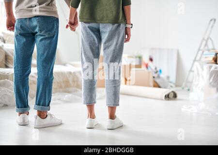 Vue arrière d'un jeune couple affectueux en Jean et pullovers tenant les mains tout en restant debout dans la salle de séjour d'une nouvelle maison ou d'un appartement Banque D'Images