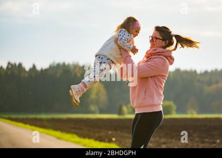 Mère tenant un enfant heureux dans l'air sur la route de campagne entre les champs agricoles contre le fond forestier. Banque D'Images