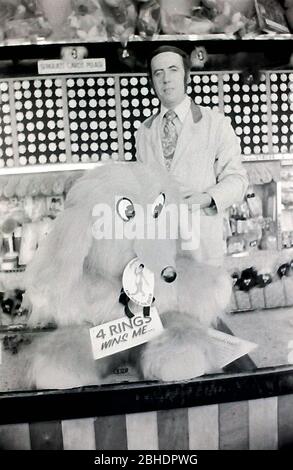 La vie quotidienne dans les rues de Blackpool, en Angleterre, en 1974. Un homme travaillant dans un stand sur la promenade de Blackpool, Lancashire, Royaume-Uni, invite les passants - en jetant des anneaux pour essayer de gagner un grand animal jouet mou. Banque D'Images