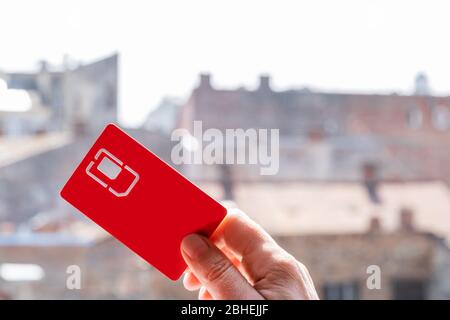 La main tient un emplacement rouge de carte SIM sur le fond de la rue avec ciel et maisons, espace de copie. Banque D'Images