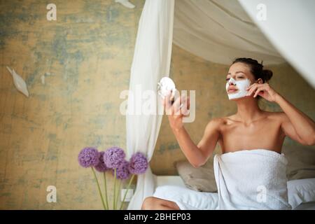 Jeune femme gaie masque facial sur le visage assis dans le lit et regardant le miroir.Beauté et concept de soins du corps. Banque D'Images