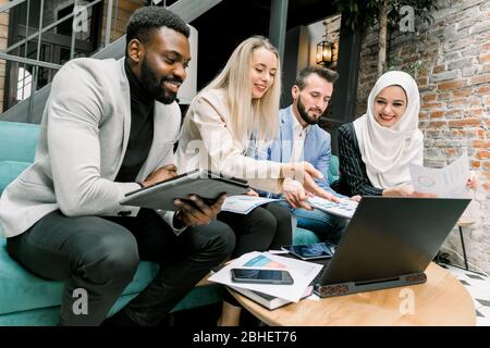 Vue latérale de quatre jeunes collègues professionnels multiethniques satisfaits, souriant tout en regardant ensemble sur l'écran d'un ordinateur portable avec de bons résultats de Banque D'Images