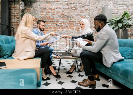 Agréable quatre jeunes gens d'affaires multiethniques, hommes et femmes, assis sur le sofa doux dans le bureau moderne près de mur décoratif en briques et Banque D'Images