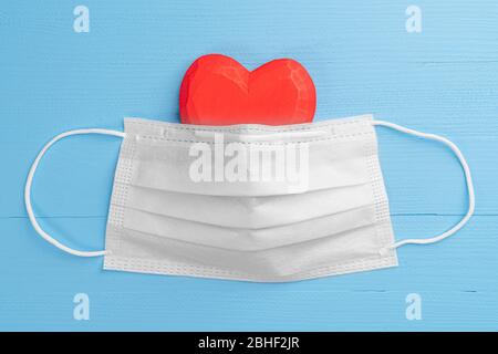 Coeur en bois rouge recouvert d'un masque de protection médical blanc sur fond bleu clair en bois. Gros plan. Concept médical Banque D'Images