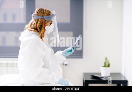 Une femme médecin assise dans une salle d'hôpital à l'aide du téléphone portable portant un costume de sécurité et un masque de protection et des gants en latex Banque D'Images