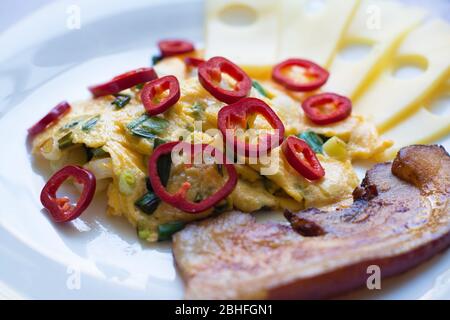 Œufs brouillés avec oignons verts coupés en dés, tranches de poivron rouge sucré, une épaisse tranche de bacon fumé poêlé et des morceaux de fromage Emmental. Banque D'Images