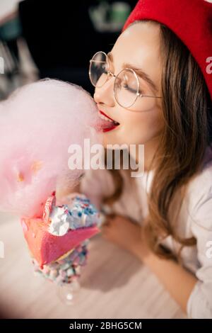 Jeune femme en béret rouge et verres a un grand dessert sucré avec des bonbons en coton rose dans un café en France Banque D'Images