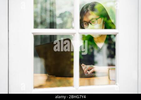 Jeune femme asiatique portant un masque et des lunettes de vue en N95 travaillant sur son ordinateur à la maison, vu par fenêtre avec reflet des arbres, pendant le verrouillage de la ville Banque D'Images
