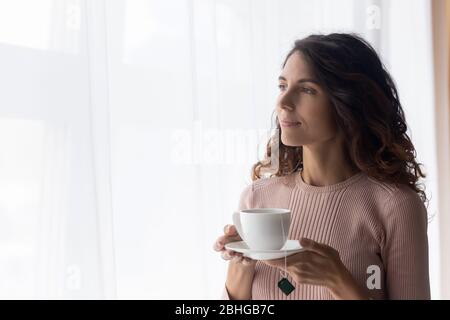 Jeune femme rêveuse se tenant près de la fenêtre avec une tasse de thé. Banque D'Images