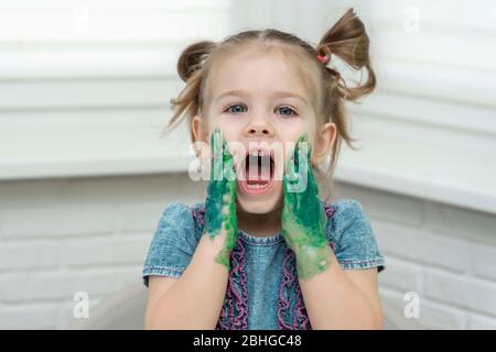 Petite fille peint avec les doigts.souriante fille touche les paumes en peinture à ses joues, auto-isolation, coronavirus covid-19, rester à la maison Banque D'Images