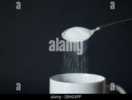 Verser du sucre blanc de la cuillère à la tasse à café blanche, tasse devant le fond noir Banque D'Images