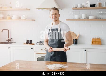 Le chef cuisinier mâle sourit le boulanger, tient la pâte fraîche dans les mains dans le tablier, cuisine légère de fond Banque D'Images