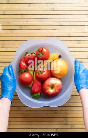 Mains avec gants en latex bleu remuant un bol bleu avec différents fruits frais et propres sur une base en bois. Bananes, tomates, pommes, fraises et Banque D'Images