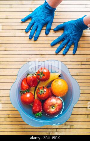 Mains avec gants en latex bleu et bol bleu avec différents fruits frais et propres sur une base en bois. Bananes, tomates, pommes, fraises et orang Banque D'Images