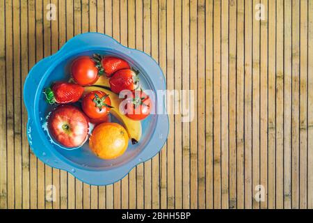 Bol bleu avec différents fruits frais et propres sur une base en bois. Bananes, tomates, pommes, fraises et oranges immergées dans l'eau avec du spac copie Banque D'Images