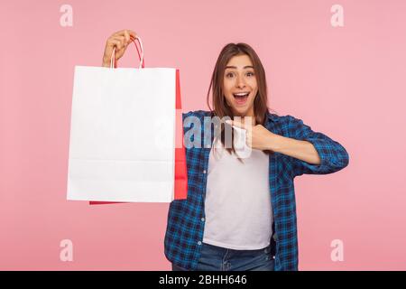 Portrait d'une jeune fille ébloui en chemise à damier regardant un appareil photo avec une expression surprise, pointant des sacs de shopping, acheter et se vanter de nouveaux produits Banque D'Images