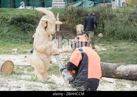 Le woodcarver en costume spécial et casque crée une sculpture en bois de mouton avec tronçonneuse. Passe-temps dans votre temps libre. Banque D'Images