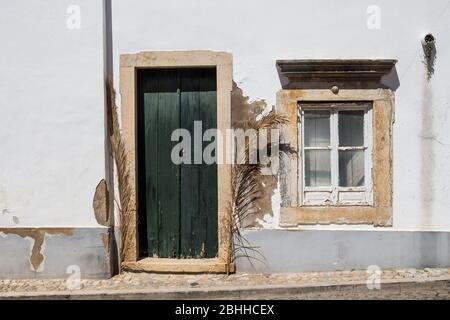 Couleur blanche de la façade d'une maison. Cadres en pierre de la porte et de la fenêtre. Porte en bois vert foncé. Fenêtre avec rideau. Séchez les branches de palmiers autour de Banque D'Images