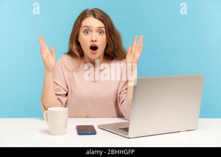 OH mon dieu, wow! Jeune femme avec une expression choquée regardant l'appareil photo avec bouche ouverte et lever les mains en surprise, travaillant sur un ordinateur portable de la maison offic Banque D'Images