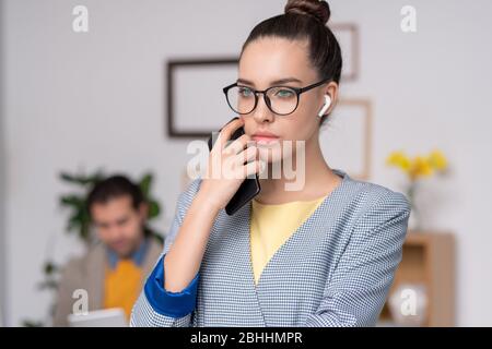Jeune femme d'affaires pensive en veste à damier bleu portant des écouteurs sans fil tenant le téléphone près du visage tout en pensant au projet Banque D'Images
