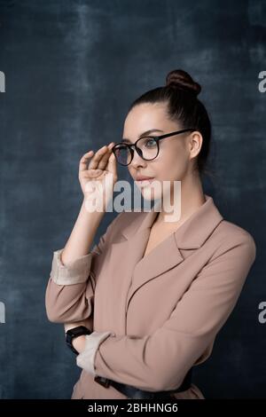 Jolie jeune femme réfléchie dans une veste élégante, se tenant contre le tableau noir et ajustant les lunettes tout en regardant de côté Banque D'Images