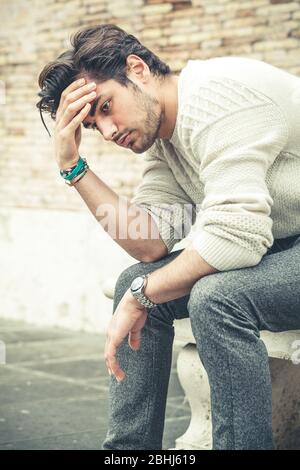 Jeune homme inquiet assis et affamé dehors avec les mains sur la tête et les cheveux avec une attitude désespérée. Pull blanc. Dans la rue. Banque D'Images