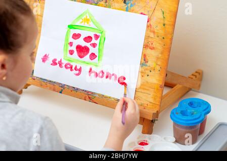 L'enfant écrit des mots rester maison avec l'aquarelle rouge sous la maison peinte avec des coeurs sur le chevalet. Concept de séjour à la maison. Banque D'Images