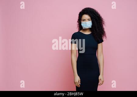 La femme afro américaine sérieuse porte un masque médical jetable sur le visage, étant sur l'auto-isolation pendant la quarantaine, reste à la maison seule, a des symptômes de cor Banque D'Images