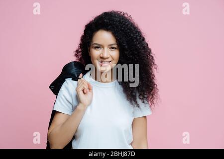 Portrait de femme ethnique positive porte un t-shirt blanc, porte la veste sur l'épaule, a une expression gaie, prêt pour pique-nique, aime jour de congé, pose dedans Banque D'Images