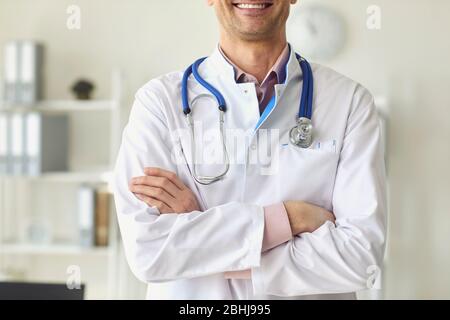 Un médecin thérapeute positif dans un manteau blanc a croisé des bras debout dans un bureau blanc d'une clinique médicale. Banque D'Images
