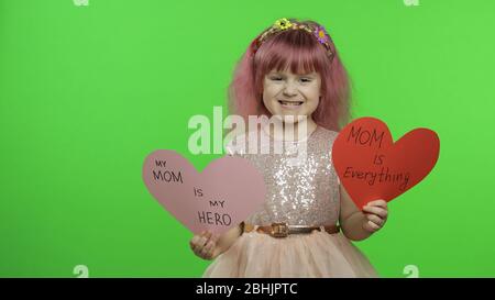 La princesse de petite fille souriante tient une coupe deux coeurs de papier avec un message texte sur maman dans ses mains. Arrière-plan Chroma Key. Un enfant mignon qui aime l'enfance heureuse, montre un signe d'amour. Concept de la fête des mères Banque D'Images