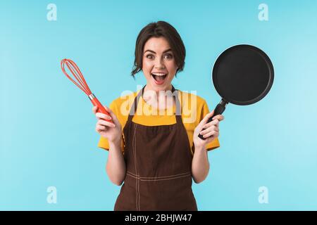 Image de jeune femme chef enthousiaste et émoussant tenant fouet et poêle isolée sur fond bleu mur. Banque D'Images