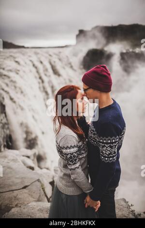 Élégant jeune couple in love kissing ensemble près de la célèbre cascade de Dettifoss paysage islandais. Des chandails de laine, chapeau, les cheveux rouges, gris jupe. Paysage spectaculaire, le temps froid en Islande. Banque D'Images