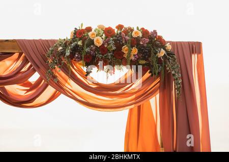 Décoration d'arche de mariage en bois lors de la cérémonie de coucher du soleil en plein air. Fleurs roses rouges et beiges, raisins et tissus suspendus. Lumière de soleil incroyable et chaude. Banque D'Images