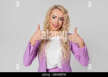 Bon travail, comme ça. Agréable belle femme blonde heureuse, montre pouce vers le haut et sourire largement, donne un avis positif, partage son opinion positive sur un mur gris Banque D'Images