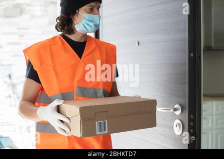 Jeune homme de livraison à la porte avant avec une boîte de colis, portant des gants et un masque de visage pendant le verrouillage de la pandémie de covid-19. Profil. Banque D'Images