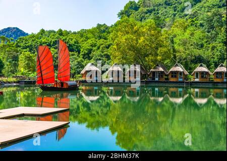 Indésirable. Magnifique bateau traditionnel vietnamien avec voiles rouges sur un lac pittoresque dans la jungle. De nombreuses petites maisons se reflètent dans l'eau. Banque D'Images