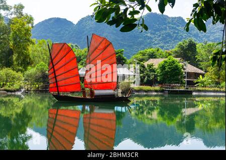 Indésirable. Magnifique bateau traditionnel vietnamien avec voiles rouges sur un lac pittoresque dans la jungle. Voiles rouges reflétées dans l'eau. Banque D'Images