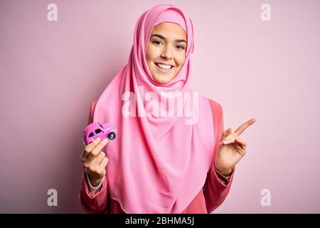 Jeune fille portant le hijab musulman tenant le petit jouet de voiture sur un fond rose isolé très heureux pointant avec la main et le doigt sur le côté Banque D'Images