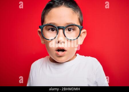 Jeune garçon intelligent enfant portant des verres d'angle sur fond rouge isolé peur en choc avec un visage surprise, peur et excité avec peur expres Banque D'Images