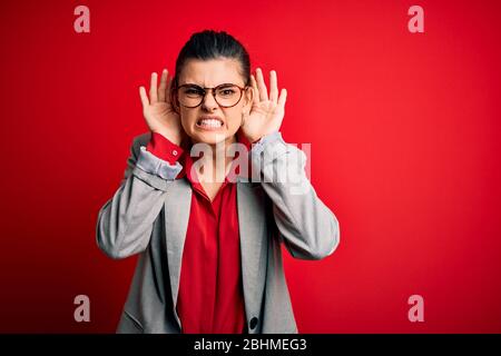 Jeune femme d'affaires brunette, portant une veste et des lunettes sur fond rouge, essayant d'entendre les deux mains sur le geste de l'oreille, curieuse pour le potins. Il Banque D'Images