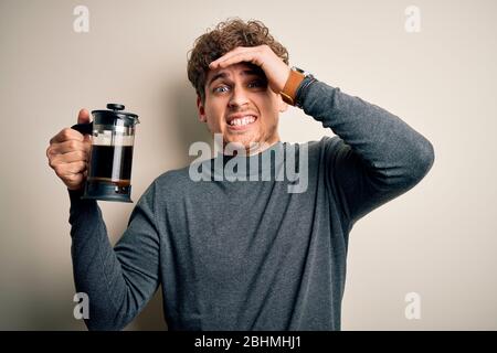 Jeune homme blond avec des cheveux frisés faisant du café à l'aide de la machine à café sur fond blanc souligné avec la main sur la tête, choqué avec la honte et le visage surprise, Banque D'Images