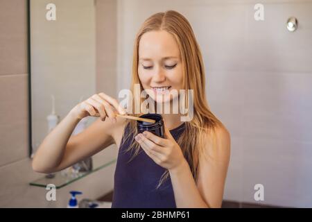 jeune femme se brossant les dents avec une pâte noire à dents avec charbon actif, et brosse noire à dents dans sa salle de bains Banque D'Images