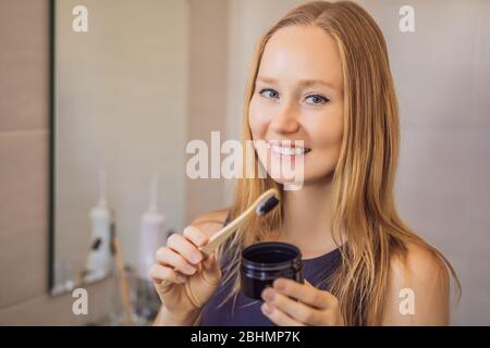 jeune femme se brossant les dents avec une pâte noire à dents avec charbon actif, et brosse noire à dents dans sa salle de bains Banque D'Images