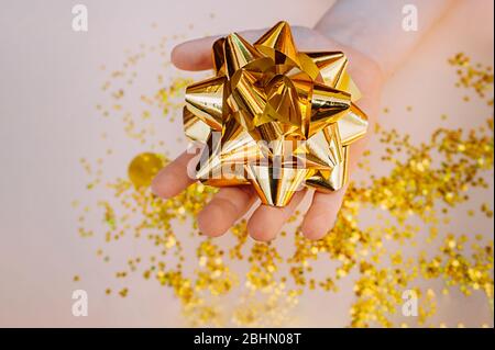 la main à peau claire tient un grand noeud en feuille d'or sur un fond d'étoiles d'or éparses sur un fond beige doux. Banque D'Images