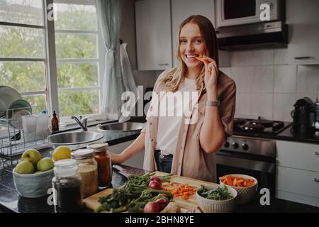 Jeune femme gaie dans la cuisine moderne préparant salade de légumes frais et manger une carotte souriant à l'appareil photo Banque D'Images