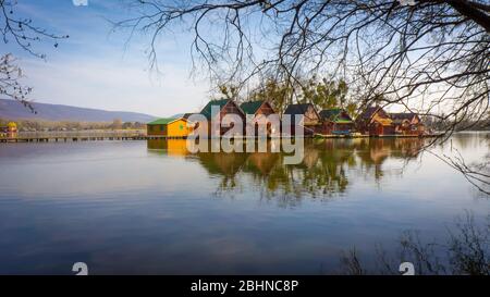 Tata, Hongrie - Petites maisons de pêcheurs en bois au lac Derito sur une île avec réflexion. L'image est entourée de branches d'arbre. Banque D'Images