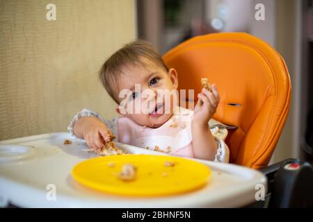 bébé mignon dans un siège d'enfant orange a incliné sa tête et regarde l'appareil photo, tenant une tarte dans ses mains. chapelure et une assiette orange sur la table. fermer- Banque D'Images