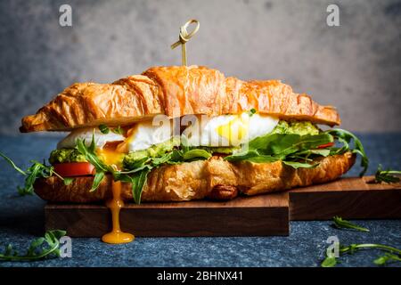 Sandwich croissant avec œufs pochés, tomates et guacamole sur un panneau en bois. Concept de restauration pour le petit déjeuner. Banque D'Images