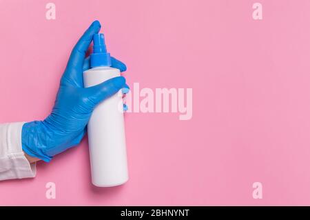 Protection contre le coronavirus. Mains dans un gant bleu jetable contenant une bouteille de désinfectant de surface ou un distributeur de désinfectant pour les mains sur fond rose. HYG Banque D'Images